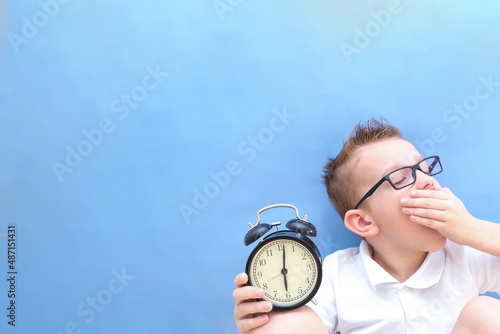 a boy on a blue background with glasses holds an alarm clock in his hands. it is difficult for a child to wake up in the morning