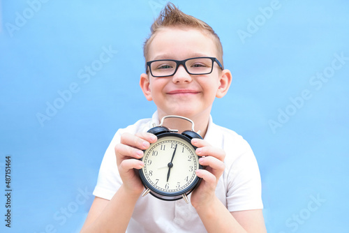a boy on a blue background with glasses holds an alarm clock in his hands. He woke up, he has a lot of energy