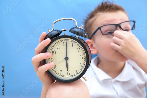 a boy on a blue background with glasses holds an alarm clock in his hands. it is difficult for a child to wake up in the morning