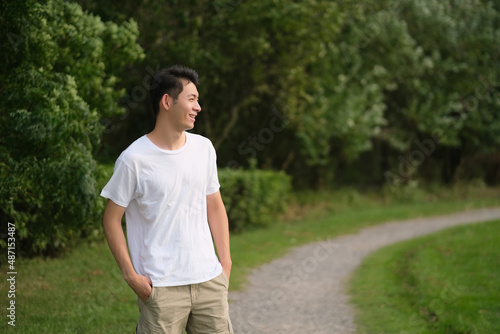 Asian young man in white T shirt smiling at green public park. Side face