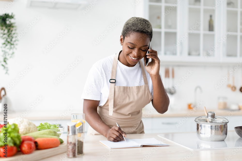 Smiling young african american woman in apron calling by phone, writing recipe on notebook at table Smiling young african american woman in apron calling by phone, writing recipe on notebook at table