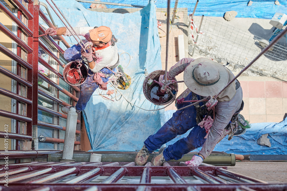 Two professional male painters work at height, hanging from ropes and a harness while painting with a brush and a bucket. Dangerous jobs, safety equipment and occupational risk prevention.