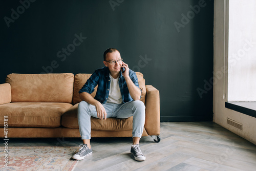 A man in casual clothes sits on the couch and talks on the phone