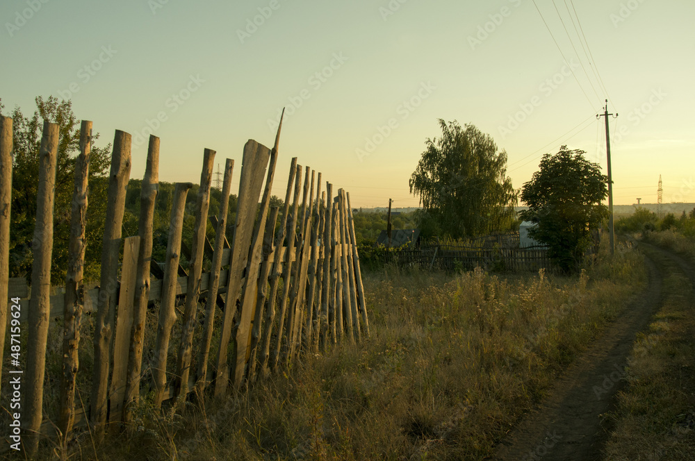 Obraz premium A dilapidated wooden fence along a rural road stretching into the distance, on a summer evening at sunset
