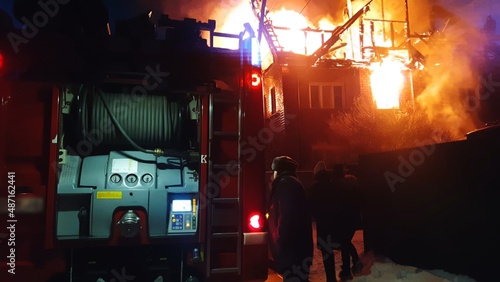 The roof of the house is on fire. The residential building burn, village. Firefighters put out a fire. Fire Department putting out flame. Fire truck, headlights and panel, siren, signal, alarm. photo