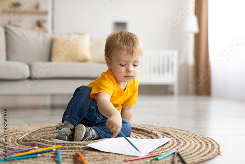 Caucasian little boy drawing picture with colorful pencils at home, sitting on floor carpet in living room interior