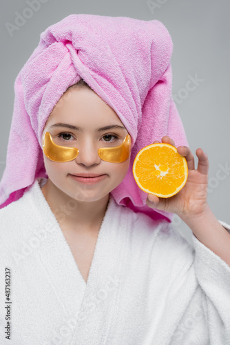 Model with down syndrome in bathrobe and eye patches holding orange and looking at camera isolated on grey