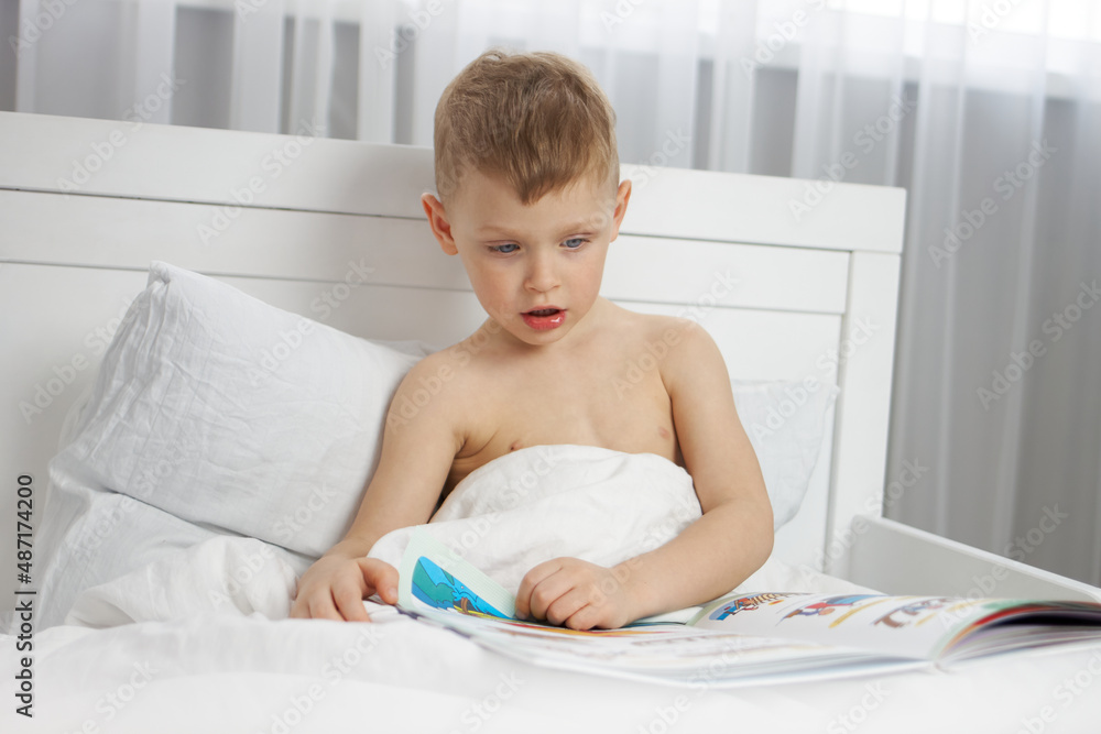 A little boy is reading a book with colorful pictures while sitting on a white bed A little boy is reading a book with colorful pictures while sitting on a white bed
