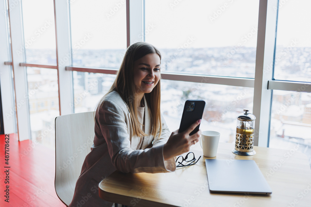 Obraz premium Cheerful young woman laughing while reading funny notification on smart phone sitting front laptop in cafe. Happy woman sending money through internet banking app on mobile phone