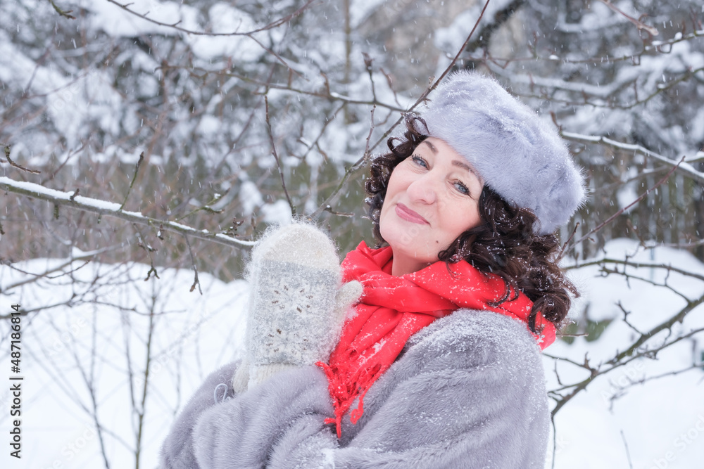 adult woman in fur coat, hat and red scarf smiling in winter in park adult woman in fur coat, hat and red scarf smiling in winter in park