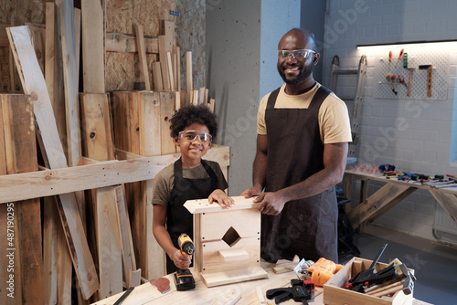 Fotografiet Portrait of happy black father building wooden birdhouse with son and looking at
