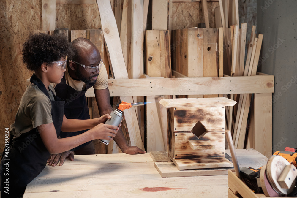 Side view portrait of African-American father and son decorating wooden birdhouse together in workshop Side view portrait of African-American father and son decorating wooden birdhouse together in workshop