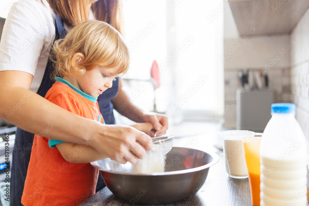 Family in a kitchen. Mother and child sieving flour into a bowl. Mom and small kid son cooking together making dough in bowl in kitchen. . Homemade food and a little helper Family in a kitchen. Mother and child sieving flour into a bowl. Mom and small kid son cooking together making dough in bowl in kitchen. . Homemade food and a little helper