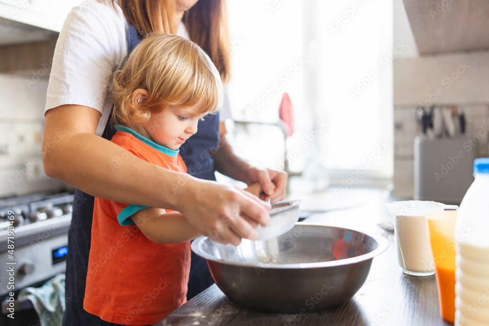 Family in a kitchen. Mother and child sieving flour into a bowl. Mom and small kid son cooking together making dough in bowl in kitchen. . Homemade food and a little helper Family in a kitchen. Mother and child sieving flour into a bowl. Mom and small kid son cooking together making dough in bowl in kitchen. . Homemade food and a little helper