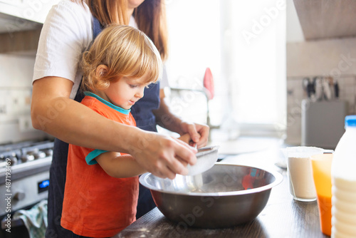 Family in a kitchen. Mother and child sieving flour into a bowl. Mom and small kid son cooking together making dough in bowl in kitchen. . Homemade food and a little helper
