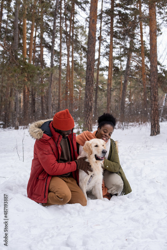 Happy African couple embracing their cute dog during their walk in the forest in winter © pressmaster