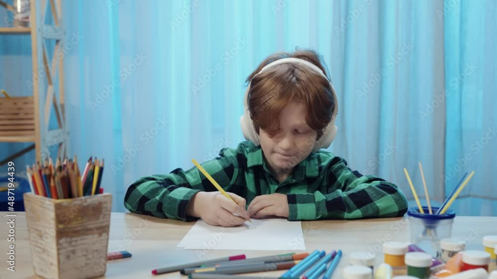 Little schoolboy sitting at table in room, drawing with pencil in album and listens to music in big white headphones. Teenager boy is engaged in creativity. Close up. Slow motion ready, 4K at 59.94fps Little schoolboy sitting at table in room, drawing with pencil in album and listens to music in big white headphones. Teenager boy is engaged in creativity. Close up. Slow motion ready, 4K at 59.94fps