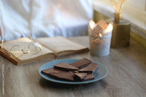 Plate of chocolate, open book, reading glasses, lit candle and flowers on the table. Selective focus.