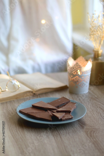 Plate of chocolate, open book, reading glasses, lit candle and flowers on the table. Selective focus.