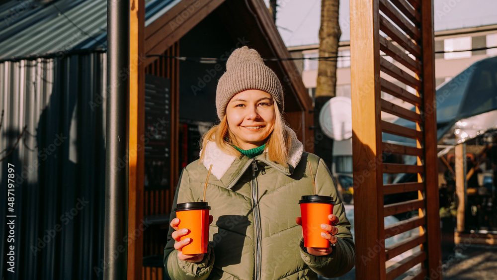 Woman carries two cups of coffee or mulled wine at the city outside food court. She looks at the camera. Street food festival, place to relax and eat Woman carries two cups of coffee or mulled wine at the city outside food court. She looks at the camera. Street food festival, place to relax and eat