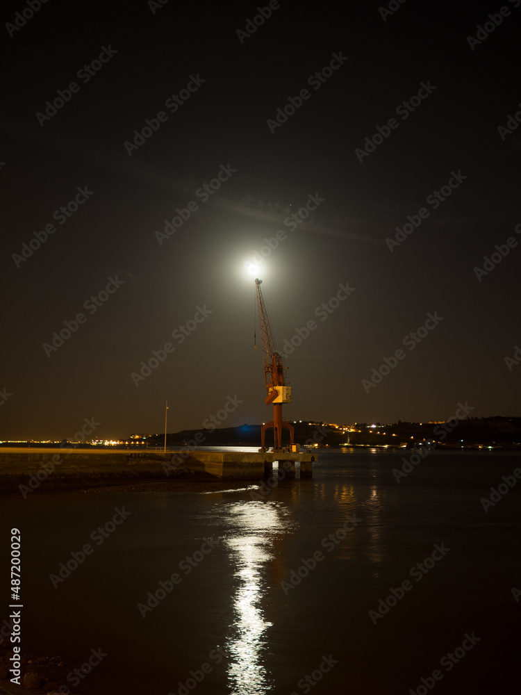 Full moon over a port crane by Lisbon Tagus river at night