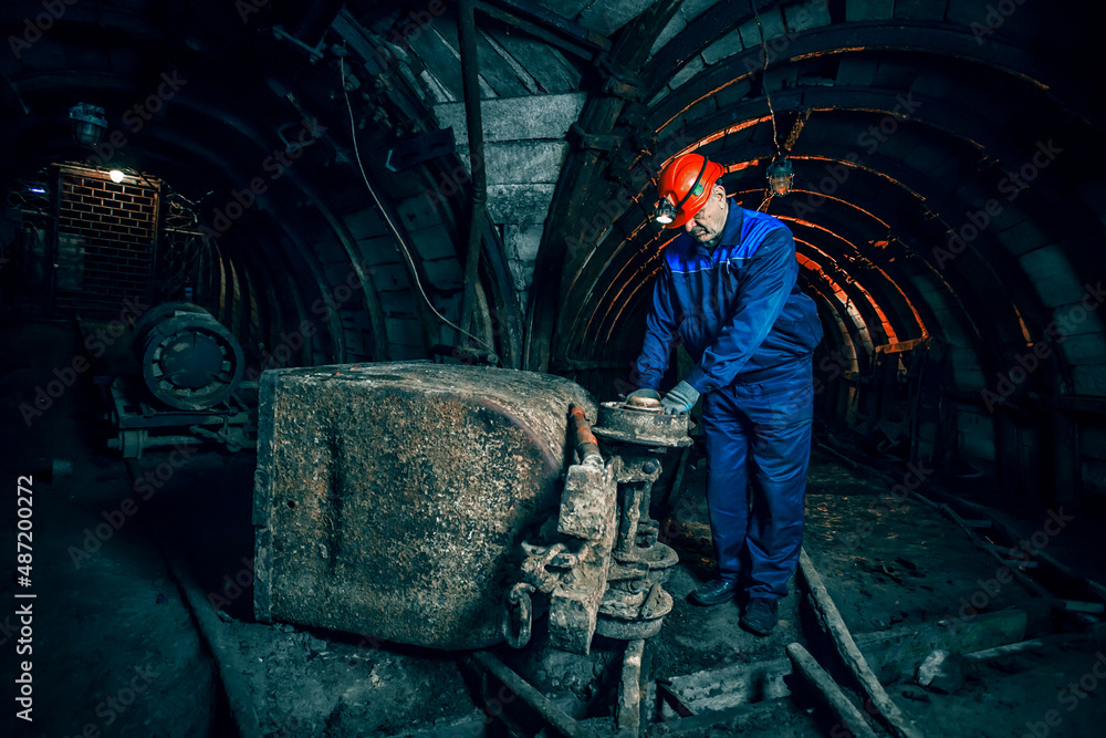 A tired miner in an old coal mine is repairing a broken trolley. A miner in a blue protective suit with an orange helmet on his head. Copy space.