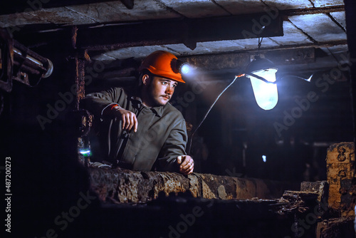 A tired young miner in an old coal mine looks at a lamp. A miner in a protective suit with a helmet on his head and a flashlight works in the dark. Work in a coal mine.