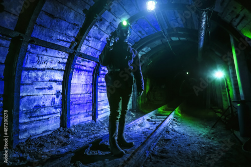 A tired young miner in an old coal mine stands with a jackhammer on his shoulder. A miner in a protective suit with a helmet on his head and a flashlight works in the dark.