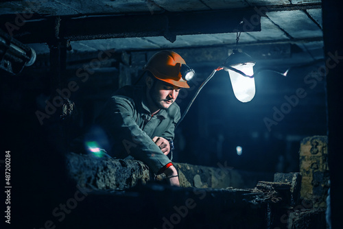 Tired young miner in an old coal mine. A miner in a protective suit with a helmet on his head and a flashlight works in the dark. Work in a coal mine.