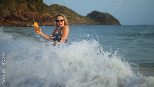 Sexy hot woman stand knee-deep in sea on golden sunset. Girl on tropical beach in green swimsuit having fun and waving his hands  drinks her orange cocktail Pina Colada. Big waves hitting buttocks