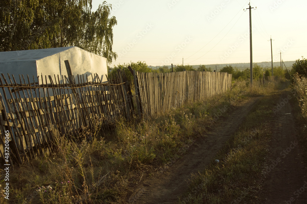 Summer rural evening landscape with a road, wooden fence, metal barn, birch, electric poles at sunset