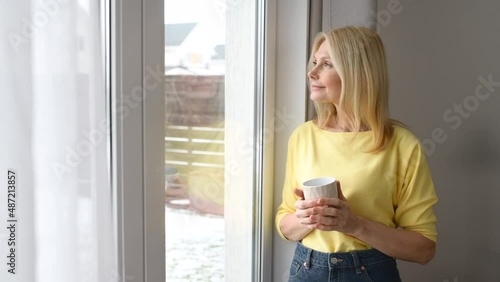 Calm and peaceful middle-aged woman takes a break, rests with a cup of hot tea near the window at home. Contented charming mature lady enjoying coffee and daydreaming