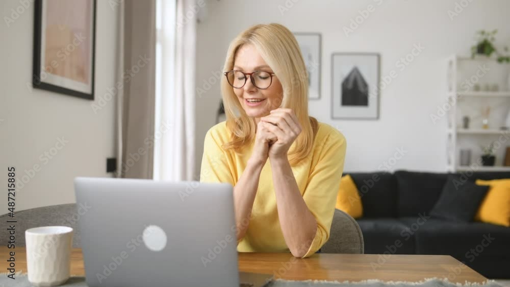 Smiling senior woman waving hello into webcam, female has video call on the laptop sitting at the desk in a home office. Video conference, online meeting, remote work Smiling senior woman waving hello into webcam, female has video call on the laptop sitting at the desk in a home office. Video conference, online meeting, remote work