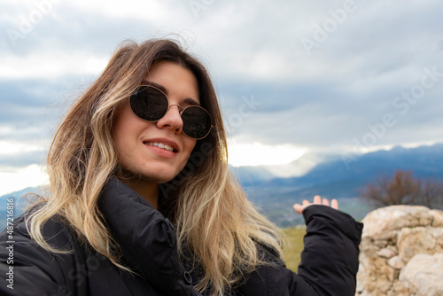 Happy beautiful woman with sun glasses takes a selfie portrait on vacation outdoor. She is standing on old archaeological site. She shows with hand and invites you. 