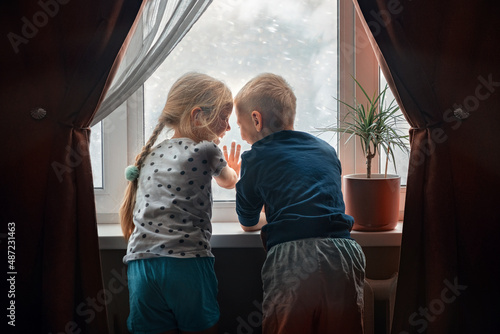 Children, a boy and a girl look out the window of their house during a snow storm