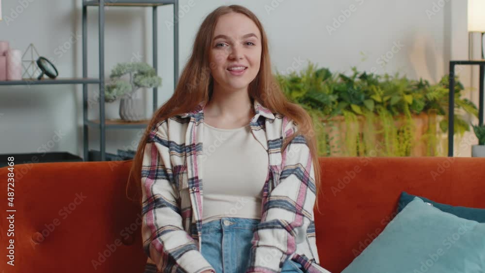 Beautiful teenager girl smiling friendly at camera and waving hands gesturing hello, hi, greeting or goodbye, welcoming with hospitable expression. Young woman indoors at home in living room on sofa Beautiful teenager girl smiling friendly at camera and waving hands gesturing hello, hi, greeting or goodbye, welcoming with hospitable expression. Young woman indoors at home in living room on sofa