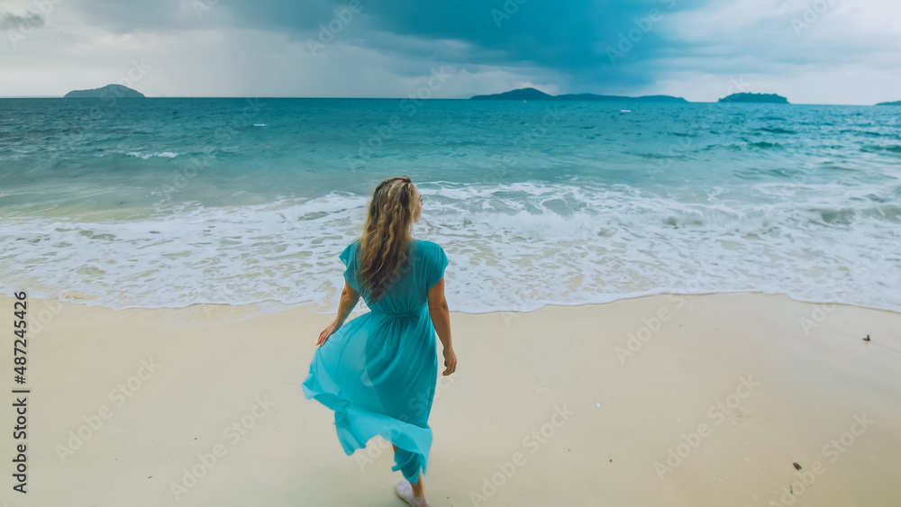 Charming woman with blonde curly long hair walk in beach, in stormy morning rain cloudy sea. Girl in turquoise swimsuit tunic. Concept resort outdoor relax summer vacation, travel. Dark dramatic view Charming woman with blonde curly long hair walk in beach, in stormy morning rain cloudy sea. Girl in turquoise swimsuit tunic. Concept resort outdoor relax summer vacation, travel. Dark dramatic view