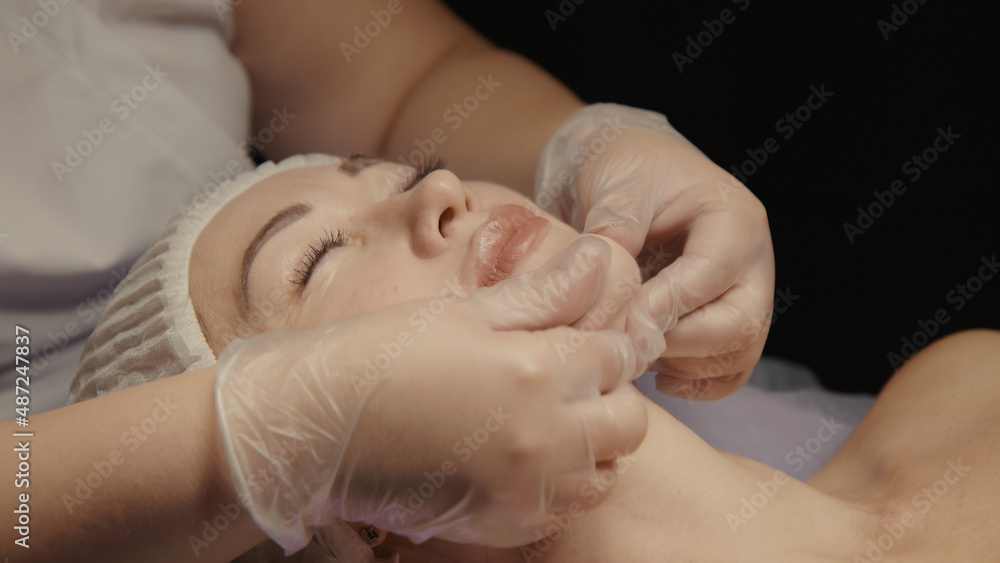 Young hypnotic face woman receiving anti-ageing facial massage in spa salon relax. Wellness body skin care face beauty treatment, rejuvenation procedure getting head face massage. Black background Young hypnotic face woman receiving anti-ageing facial massage in spa salon relax. Wellness body skin care face beauty treatment, rejuvenation procedure getting head face massage. Black background
