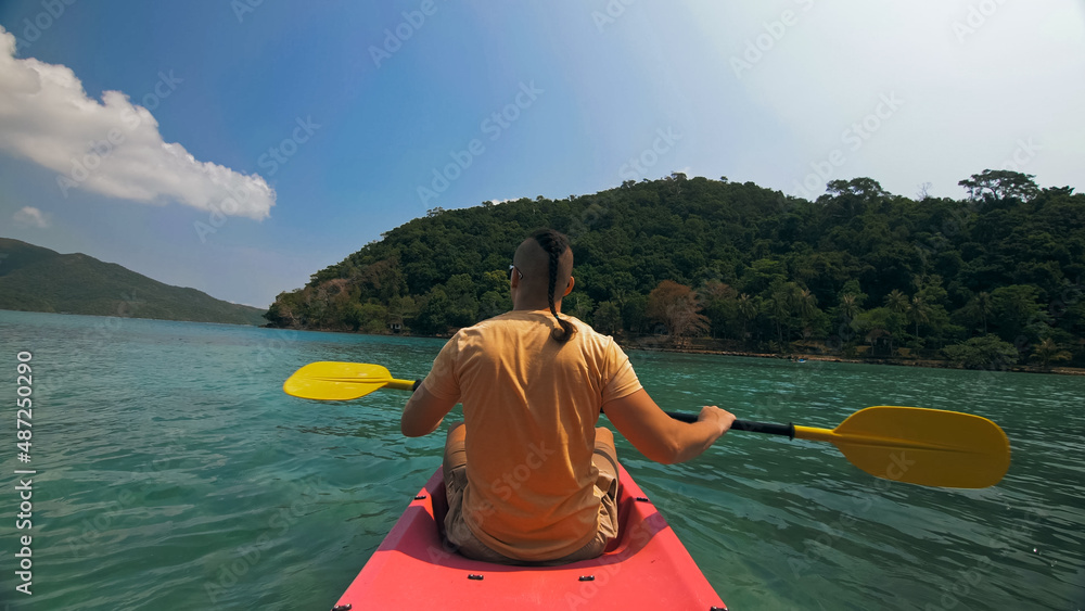 Young man with sunglasses and hat rows pink plastic canoe along sea against green hilly islands with wild jungles. Traveling to tropical countries. Strong guy is sailing on kayak in ocean, back view.