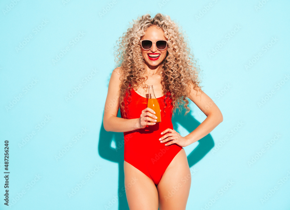 Young beautiful smiling woman posing near blue wall in studio.Sexy model in red swimwear bathing suit.Positive female with afro curls hairstyle. Happy and cheerful. Drinking lemonade from bottle Young beautiful smiling woman posing near blue wall in studio.Sexy model in red swimwear bathing suit.Positive female with afro curls hairstyle. Happy and cheerful. Drinking lemonade from bottle
