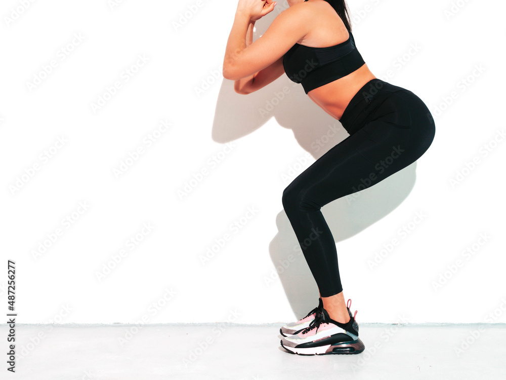 Fitness confident woman in black sports clothing. Sexy young beautiful model with perfect body. Female isolated on white wall in studio. Stretching out before training.Making squats. No face