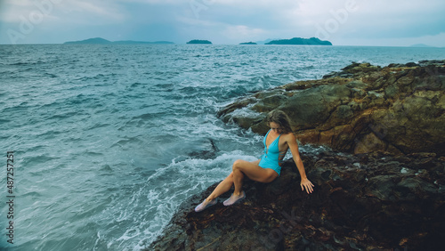 Fototapeta Naklejka Na Ścianę i Meble -  Alone attractive woman walk near a rock reef hill in stormy morning rain cloudy sea. Girl in turquoise swimsuit. Concept resort outdoor relax, vacation, loneliness. Dark dramatic view