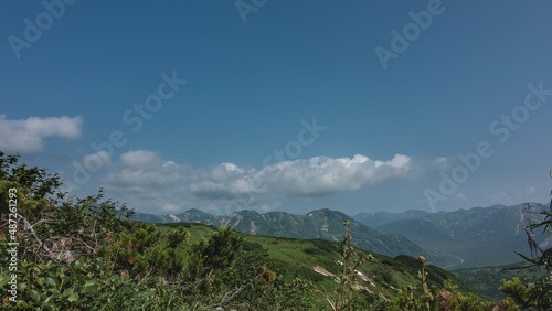 Picturesque mountain ranges against the blue sky. Clouds float over the peaks. Green vegetation in the foreground. Copy space. Kamchatka