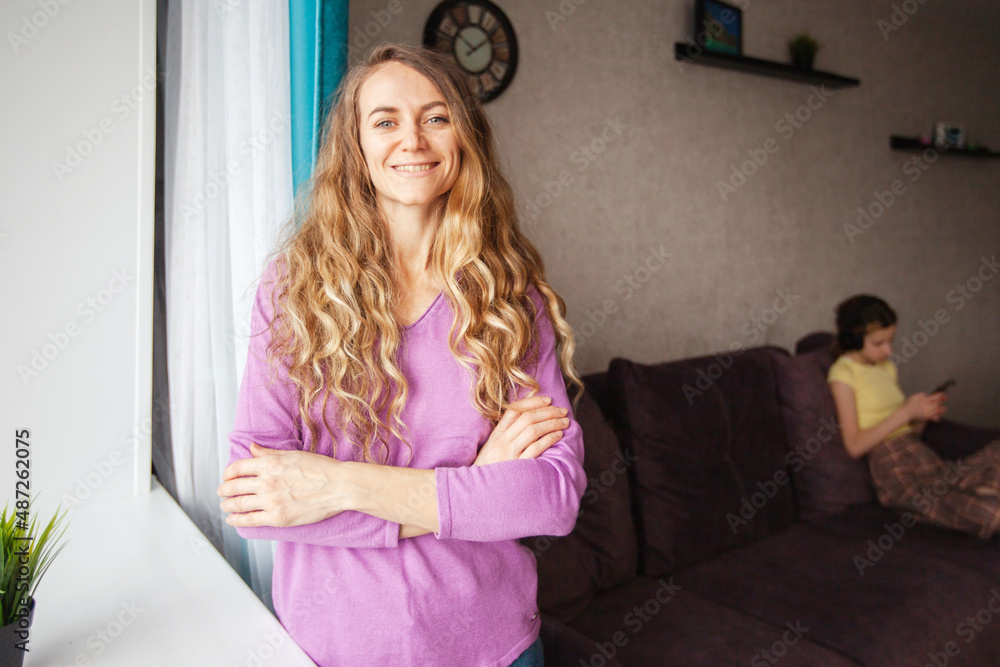 a young woman is standing at the window of the house, and in the background a girl with a phone is sitting on the couch a young woman is standing at the window of the house, and in the background a girl with a phone is sitting on the couch