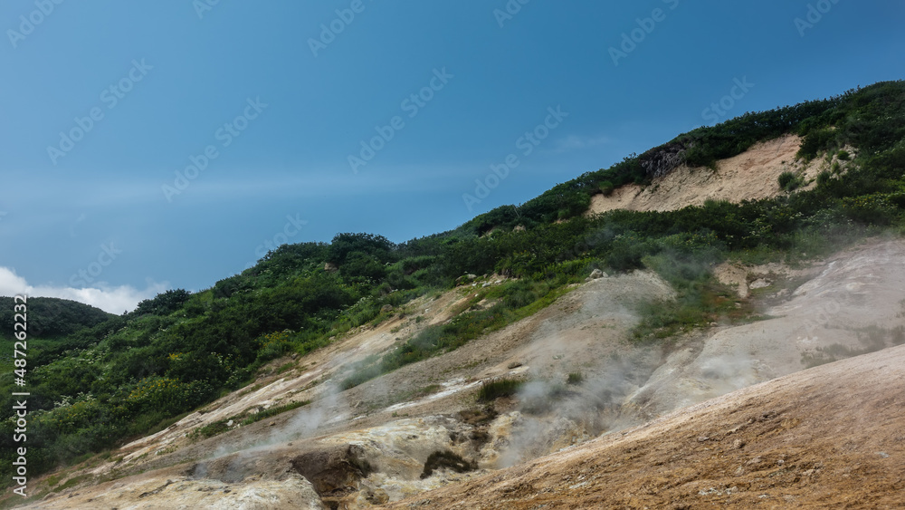 Smoke and steam from fumaroles rise above the mountain slopes. Orange sulphurous deposits on the soil. Green vegetation on the hills. Blue sky. Kamchatka
