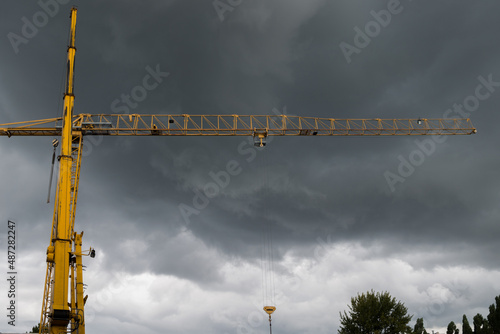 Extended telescopic jib of crane on left. Behind it reinforced post of yellow tower crane with horizontal boom with hook holder moved to center with hook hangs on chains on background of stormy sky.