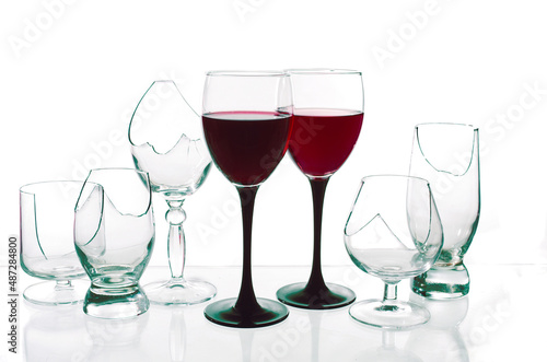 Two glasses with red wine surrounded by broken glasses on a white background.