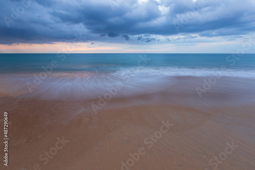 The view of the beach has white foam waves hitting the sand with turquoise water. Fluid dynamics, wave action in low light at sunset