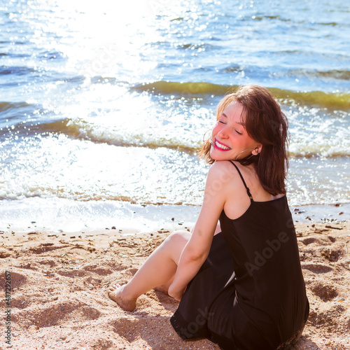 Fototapeta Naklejka Na Ścianę i Meble -  Happy Excited Caucasian Brunette Girl in Casual Clothing Relaxing on Sandy Beach In Front of Sea Shore At Daytime.
