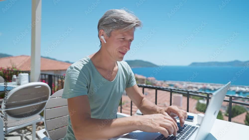 middle aged man specialist wearing in wireless earphones headset holding video call on laptop, remote work conference. man manager or freelancer working during vacation on sea resort. middle aged man specialist wearing in wireless earphones headset holding video call on laptop, remote work conference. man manager or freelancer working during vacation on sea resort.